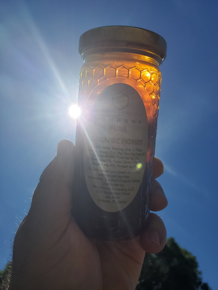 Hand holding a jar of Sting Organic Honey up with the sun peaking out from behind the bottle with a clear blue sky background